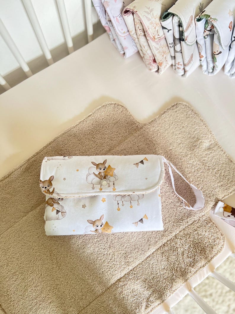 May include: A white changing pad with a cute deer and star print. The pad has a brown strap and is folded in half. It is sitting on a brown, fluffy changing mat.