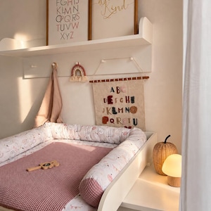 Könnte beinhalten: Eine Wickelkommode mit einer rosa Decke, dekorativen Kissen und einem Holzspielzeug. Darüber zeigen gerahmte Kunstwerke das Alphabet und die Worte „Be Kind“. Eine kleine Regenbogen-Dekoration und ein Stoffbanner mit Buchstaben sind ebenfalls zu sehen.