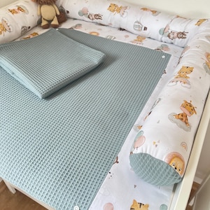 Könnte beinhalten: Ein Wickeltisch mit einer hellblauen Waffel-Wickelauflage und einem passenden Kissen. Der Tisch ist von einem Nestchen mit weißem Hintergrund und einem sich wiederholenden Muster aus Zeichentricktigern, Löwen und Ballons umgeben. Ein Stofftier-Löwe sitzt auf dem Wickeltisch.