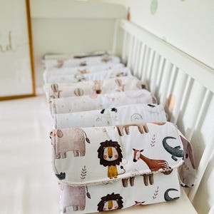May include: A stack of white changing pads with a brown, white, and beige animal print. The print features a lion, elephant, giraffe, and crocodile. The changing pads are folded and stacked on a white crib.