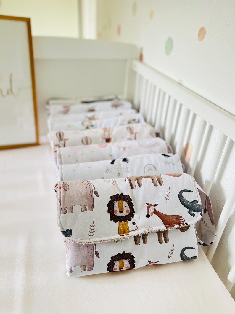 May include: A stack of white changing pads with a brown, white, and beige animal print. The print features a lion, elephant, giraffe, and crocodile. The changing pads are folded and stacked on a white crib.