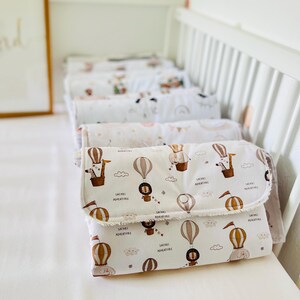 May include: A stack of white changing pads with a safari animal print and the text "Safari Adventure". The pads have a brown, white, and beige color scheme and feature illustrations of hot air balloons, giraffes, lions, and elephants.