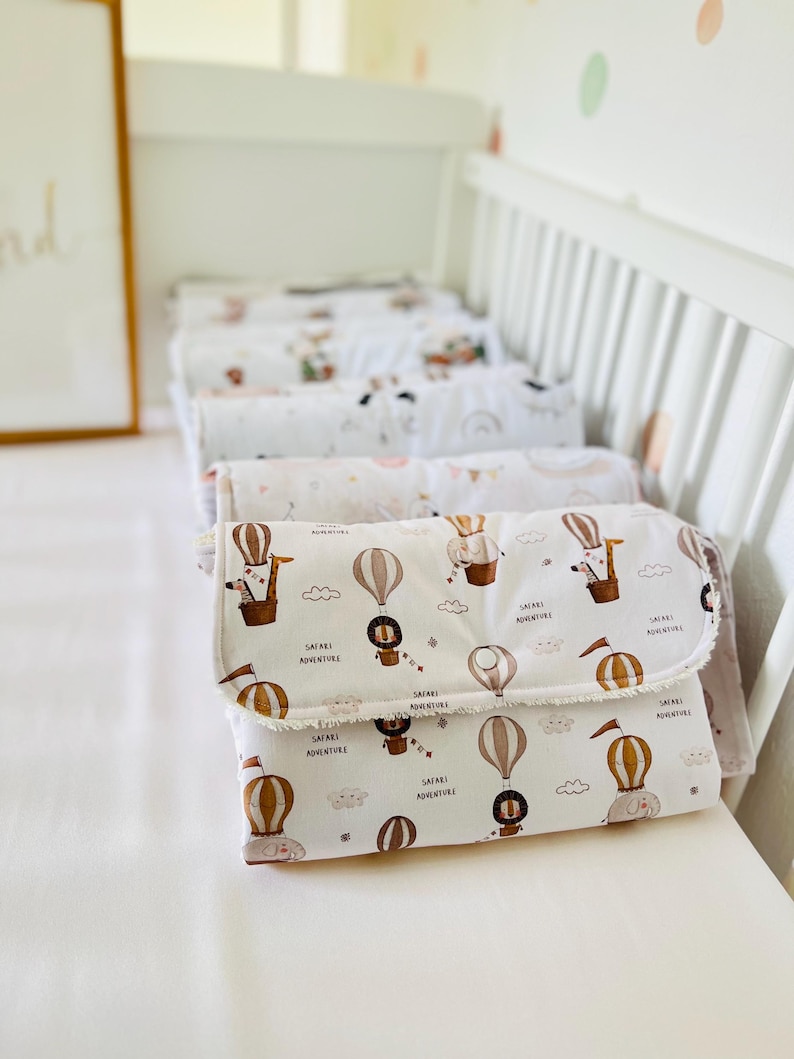 May include: A stack of white changing pads with a safari animal print and the text "Safari Adventure". The pads have a brown, white, and beige color scheme and feature illustrations of hot air balloons, giraffes, lions, and elephants.