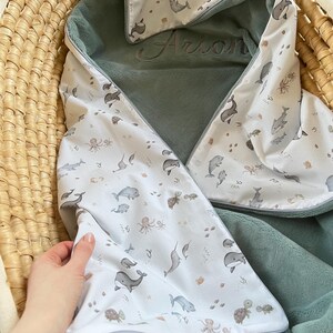 Könnte beinhalten: Eine Kapuzen-Babydecke mit Meeresmotiven. Die Decke hat ein weißes Muster mit Illustrationen von Walen, Delfinen und anderen Meeresbewohnern. Die Innenseite ist in einem weichen Salbeigrün gehalten. Der Name "Arion" ist auf der Kapuze gestickt.
