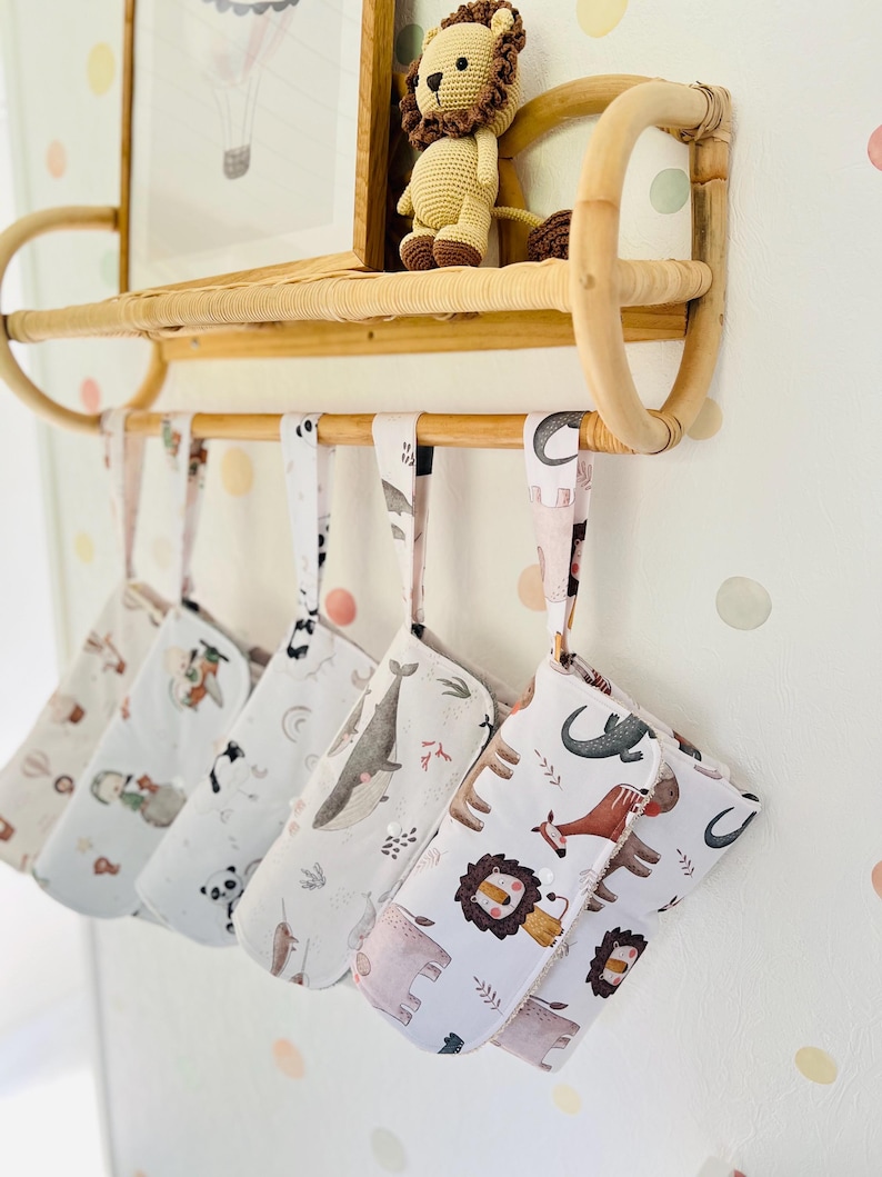 May include: Five white fabric diaper clutch bags hanging on a bamboo shelf. Each bag has a different animal print design, including a lion, elephant, whale, and other animals. The bags have a white background with brown, pink, and orange accents.