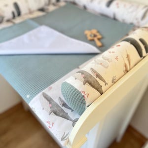 Puede incluir: Una mesa cambiador blanca con un cambiador azul y un paño blanco. La mesa tiene un protector de cuna con temática de ballenas y un juguete de madera. El protector y el cambiador tienen un diseño de ballenas y corales.