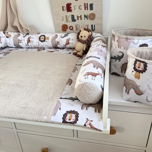 Può includere: Un fasciatoio per bambini con un cassettone bianco, un fasciatoio e un paracolpi a tema safari. Il paracolpi e la fodera del fasciatoio presentano illustrazioni di animali. Un piccolo leone di peluche è seduto sul fasciatoio. Sono visibili due cesti portaoggetti coordinati.