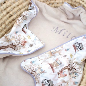 Könnte beinhalten: Eine weiße und beige Babydecke mit einem Waldtier-Druck und einem hellvioletten Saum. Die Decke ist mit dem Namen "Milo" in grauer Stickerei personalisiert.