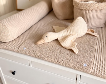May include: A beige waffle-textured changing pad cover with a matching bolster, two storage baskets, and a soft goose-shaped plush toy. The changing pad features white snap buttons and a textured fabric surface, resting on a white dresser with black knobs.