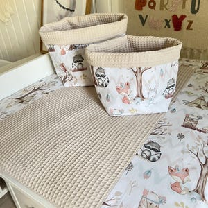 Op de afbeelding: Twee beige opbergmanden van wafelstof met een voering met een bosdierprint. De manden staan op een beige wafelstof verschoningsmat met een bosdierprintontwerp. De verschoningsmat staat op een witte commode met een lade.