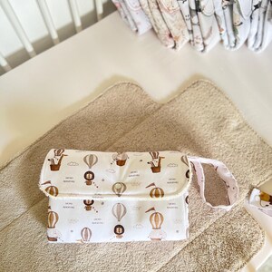 May include: A white changing pad with a safari animal print and the words "Safari Adventure" repeated. The pad has a brown strap and is folded in half. It is sitting on a beige changing mat.