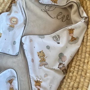 Può includere: Una coperta per neonati bianca con bordi grigi, con animali dei cartoni animati, palloncini e arcobaleni. La coperta ha un interno morbido e felpato e il nome "Leve" ricamato in grigio.