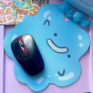 May include: A light blue, cloud-shaped mousepad with a smiling face design. A black and red computer mouse rests on the mousepad. Several blue dice are visible in the background. The mousepad is a fun accessory for a computer setup.