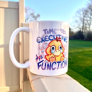 May include: White ceramic mug with a handle, featuring a cartoon duck with tears and the text "Time to Executive Dys Function" in purple and blue. The duck is yellow with orange feet and a red crest.