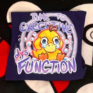 May include: A navy blue t-shirt featuring a cartoon duck with tears, surrounded by bubbles and the text "TIME TO EXECUTIVE FUNCTION". The words "dys" and "function" are crossed out. The design is in shades of purple, yellow, and blue.