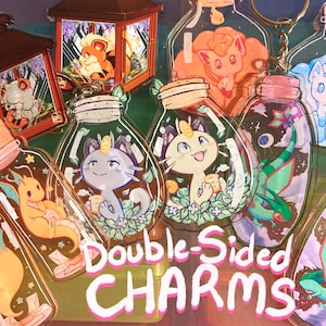 May include: Double-sided charms featuring various Pokemon characters in glass bottles. The charms are colorful and feature a variety of designs, including a blue cat, a yellow cat, a pink Eevee, a green dragon, and a yellow dragon.