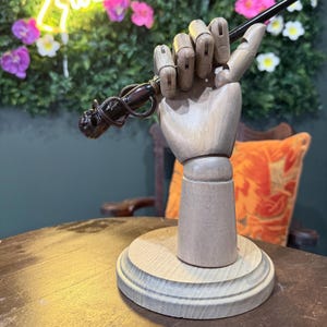 May include: A wooden articulated hand holds a dark wand with a skull design. The hand is mounted on a wooden base. The background includes flowers and a decorative pillow.