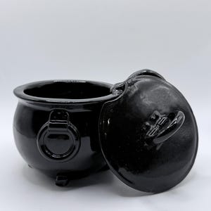 May include: A glossy black cauldron with a matching lid. The cauldron has a rounded body, three small feet, and a ring handle on each side. The lid is slightly ajar, revealing the interior.