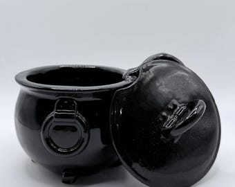 May include: A glossy black cauldron with a matching lid. The cauldron has a rounded body, three small feet, and a ring handle on each side. The lid is slightly ajar, revealing the interior.