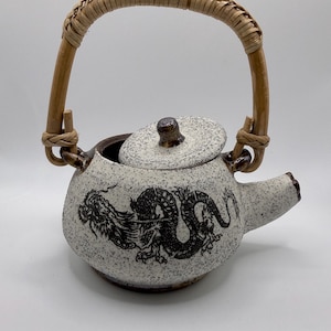 May include: Speckled ceramic teapot with a dragon design. The teapot has a woven handle and a lid with a small knob. The spout is on the right side. The dragon is black, and the teapot is white with brown speckles.