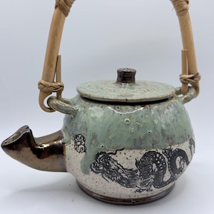 May include: A ceramic teapot with a light green and white glaze, featuring a black dragon design. The teapot has a bamboo handle wrapped in woven material and a bronze-colored spout. The lid is speckled with a dark brown knob.