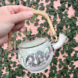 May include: A ceramic teapot with a speckled white finish and a black dragon illustration. The pot features a light green glaze drip pattern, a matching lid with a small knob, and a curved, light-colored woven rattan handle attached to two dark metal loops.