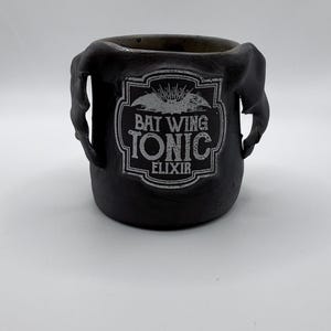 May include: A black ceramic cauldron-shaped container with two handles. The container has a white label with the words "BAT WING TONIC ELIXIR" in a gothic font. The container is on a white background.