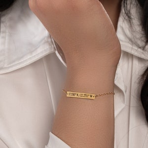 Puede incluir: Pulsera dorada con una barra rectangular grabada con coordenadas: 37.5585° N, 122.2711° W. La pulsera es delicada y está unida a una fina cadena.