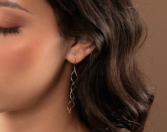 May include: A woman with long brown hair wearing a pair of long, thin, gold earrings with a twisted design.