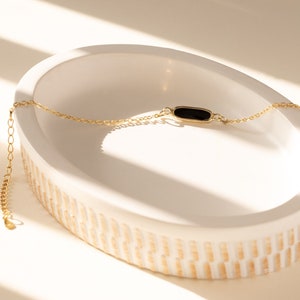 May include: A delicate gold-toned bracelet with a black rectangular centerpiece rests in a white, oval-shaped dish. The dish has a textured, ridged pattern around the base. The bracelet is displayed in natural light, creating shadows.