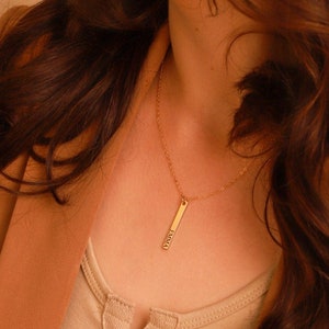 Op de afbeelding: Een gouden ketting met een verticale staafhanger met daarop "LOVE". De ketting wordt gedragen door een persoon met lang bruin haar.