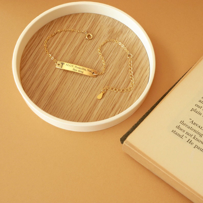 May include: A gold-colored bracelet with a rectangular pendant. The pendant has the text "ALGY: Penicillin, Nut, Bee sting" engraved on it. The bracelet is on a wooden surface inside a white tray.