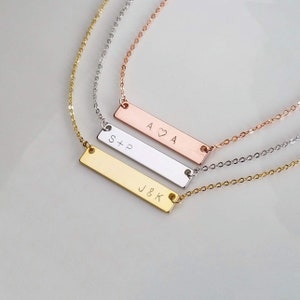 Peut inclure: Trois colliers personnalisés avec des barres en or, argent et or rose. Chaque collier présente un pendentif rectangulaire gravé d'initiales et une chaîne délicate. Les pendentifs affichent les initiales "J&K", "S+P" et "A♡A".