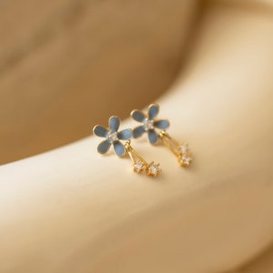 Może przedstawiać: Para złotych kolczyków wiszących z niebieskimi kwiatowymi zawieszkami z emalii i przezroczystymi kryształowymi akcentami.