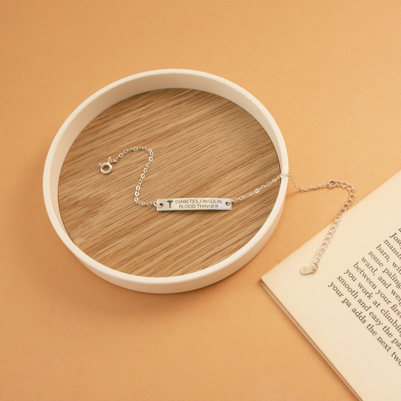 May include: A silver-colored medical alert bracelet with the text "DIABETES / INSULIN / BLOOD THINNER" engraved on a rectangular bar. The bracelet is on a wooden surface inside a white circular tray, with a book partially visible in the background.