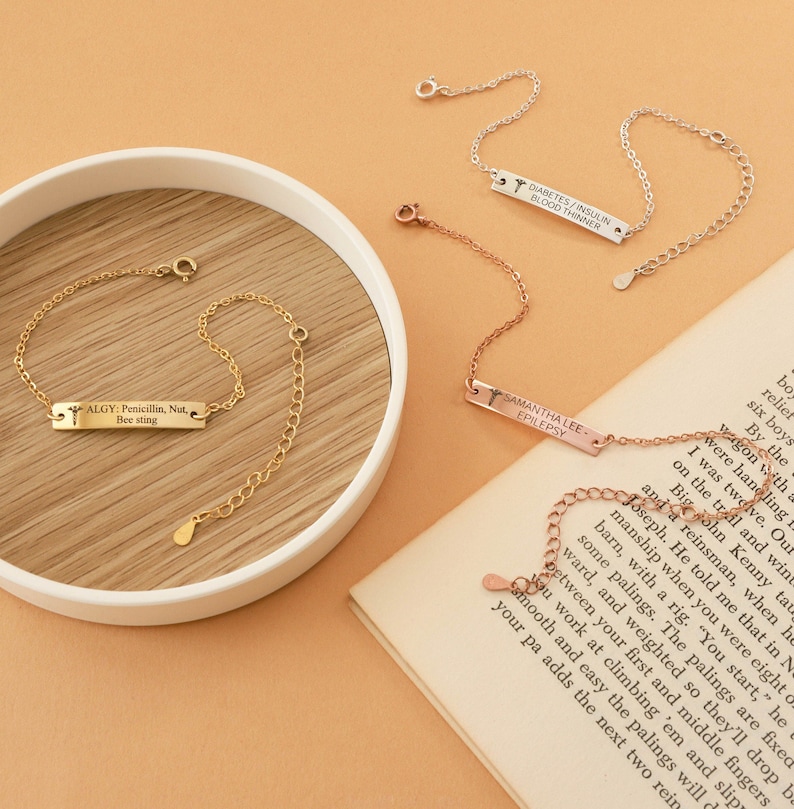May include: Several delicate medical alert bracelets in gold, silver, and rose gold. Each bracelet features a rectangular tag with engraved text, including medical conditions and names. The bracelets are displayed on a neutral background.
