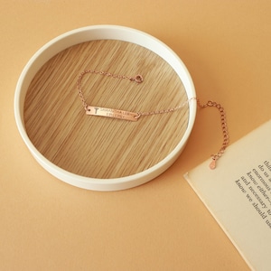May include: Rose gold bracelet with a rectangular bar engraved with "SAMANTHA LEE - EPILEPSY" and a medical symbol. The bracelet is displayed on a wooden surface inside a white, round tray. The background is a solid, light orange color.