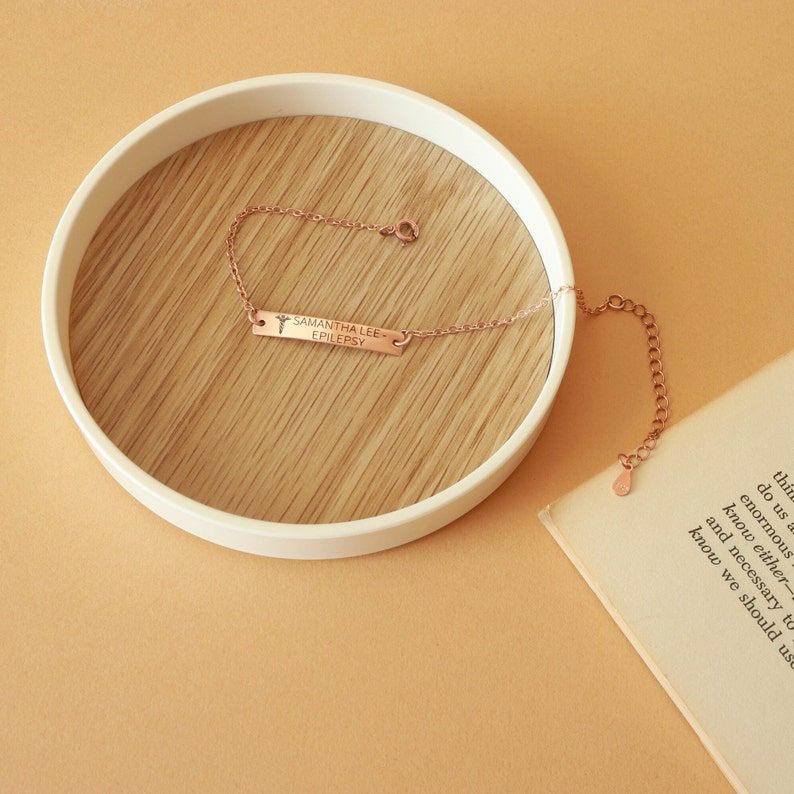May include: Rose gold bracelet with a rectangular bar engraved with "SAMANTHA LEE - EPILEPSY" and a medical symbol. The bracelet is displayed on a wooden surface inside a white, round tray. The background is a solid, light orange color.