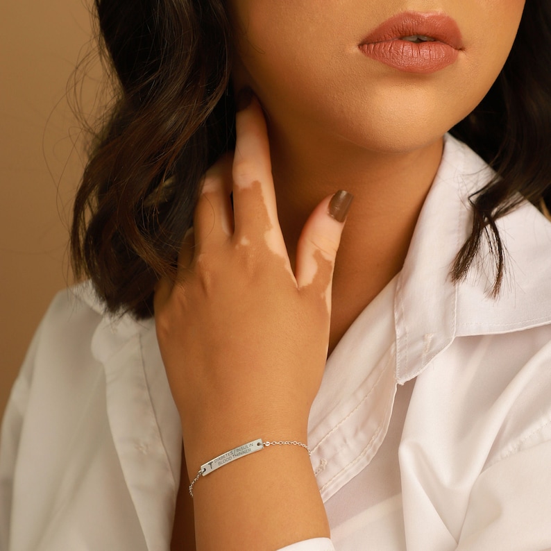 May include: A silver bracelet with a rectangular charm. The charm has engraved text and a medical symbol. The bracelet is on a wrist, and the person is wearing a white shirt.