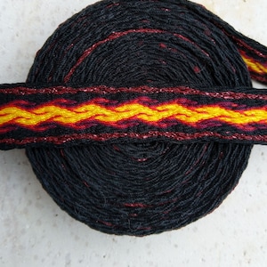 Può includere: Una bobina arrotolata di passamaneria nera tessuta con un vivace motivo a fiamma in giallo, arancione e rosso. La passamaneria ha un accento di filo metallico. Il testo "Astridas Atelier" è in basso.