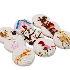 Könnte beinhalten: Ein Set aus zehn Stoffknöpfen mit Waldtieren und -szenen. Die Knöpfe sind weiß mit bunten Illustrationen eines Eichhörnchens, eines Rehs, eines Kaninchens, eines Fuchses, eines Mädchens in einer roten Kapuze und eines Baumes mit Blättern.