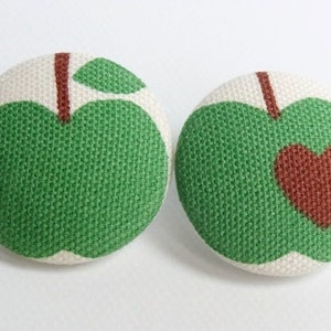 Könnte beinhalten: Zwei grüne Stoffknöpfe in Apfelform mit weißen Mittelpunkten. Ein Knopf hat ein braunes Herz in der Mitte des Apfels.