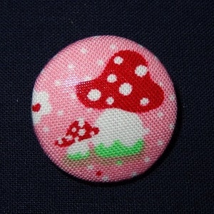 May include: A pink fabric button featuring a red and white mushroom design. The mushroom has white polka dots and a green base.