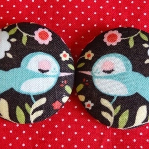 Pode incluir: Dois botões redondos de tecido com fundo preto e um pássaro azul com detalhes brancos e rosados. Os botões apresentam um design floral com flores vermelhas, brancas e amarelas.
