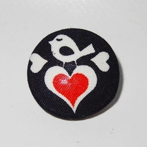 Könnte beinhalten: Ein schwarzer Stoffknopf mit einem weißen Vogeldesign, der ein rotes Herz in seinem Schnabel hält. Der Vogel sitzt auf einem weißen Knochen mit einer Herzform am Ende.
