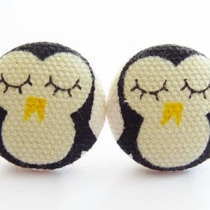 Może przedstawiać: Dwa białe guziki z tkaniny z czarnym i żółtym haftem przedstawiającym śpiącego pingwina.