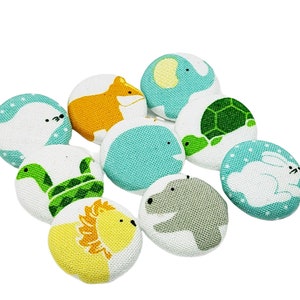 Könnte beinhalten: Ein Set aus neun Stoffknöpfen mit verschiedenen Tiermotiven, darunter eine Schildkröte, ein Flusspferd, ein Löwe, ein Fuchs, ein Kaninchen, eine Schlange, eine Robbe, ein Elefant und ein Wal. Die Knöpfe sind alle rund und haben einen weißen Hintergrund mit bunten Tierillustrationen.