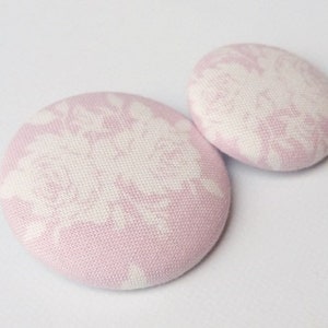 Può includere: Due bottoni in tessuto rosa con un motivo floreale bianco.
