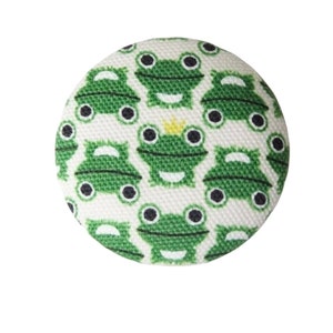 Pode incluir: Um botão branco com um padrão de sapos verdes. Os sapos têm olhos pretos e uma coroa amarela em um dos sapos.