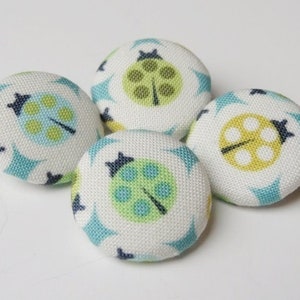 Könnte beinhalten: Vier weiße Stoffknöpfe mit einem grünen, blauen und gelben Marienkäfer-Design.